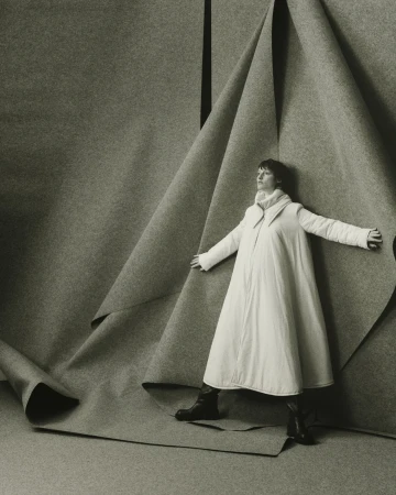 Monochrome fashion shot of an adult woman in a white coat, posing against folded gray fabric backdrop in a photo studio.