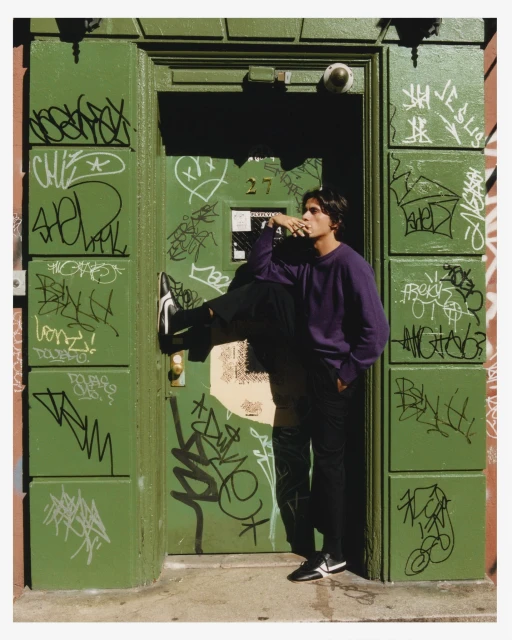 An adult posing in front of a green door covered in graffiti, with a dark purple long-sleeve shirt and dark pants and black sneakers.