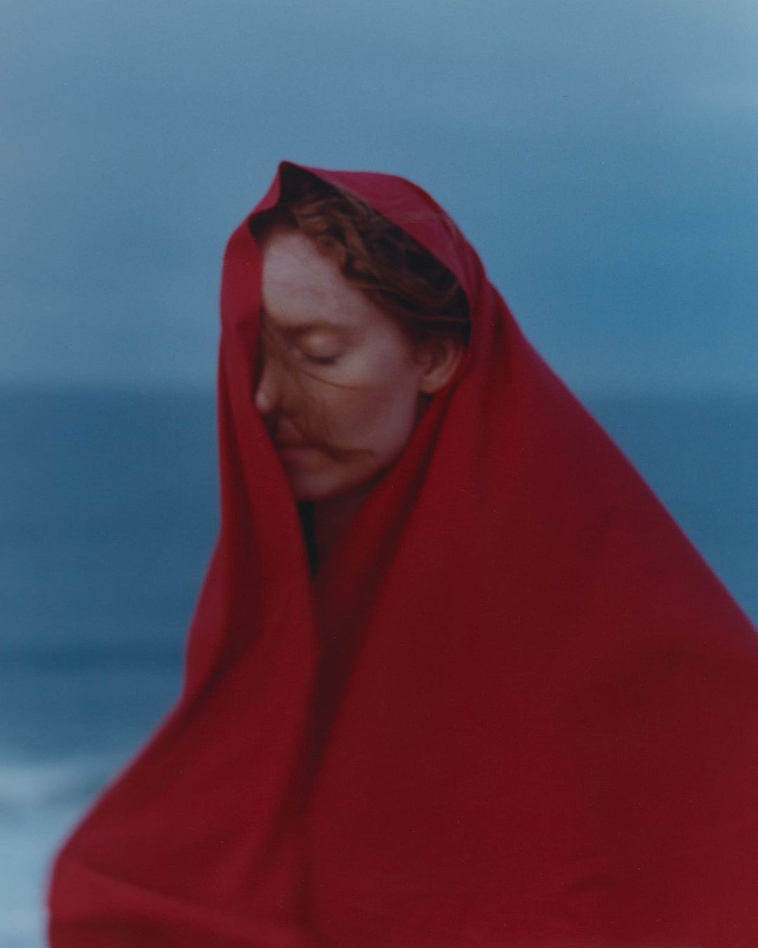 Woman in a red fabric wrap stands at the beach. Calm ocean and overcast sky in the background. Moody, evocative fine art photograph.