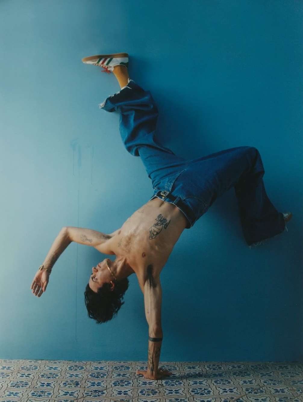 Athletic adult does a handstand, wearing jeans and sneakers against a blue wall with patterned tile floor in a fitness pose.