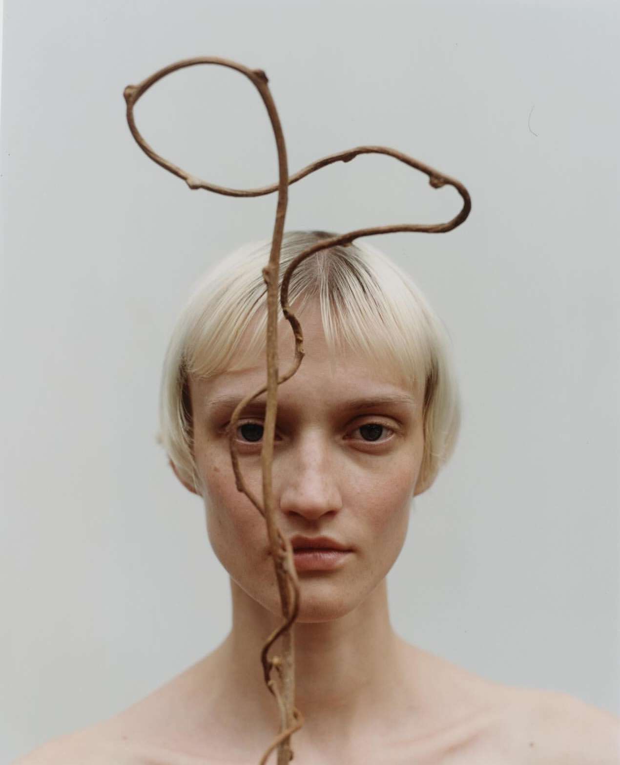 Portrait of a fair-skinned woman with short blonde hair, posing with a curly brown vine against a light grey background in studio lighting.