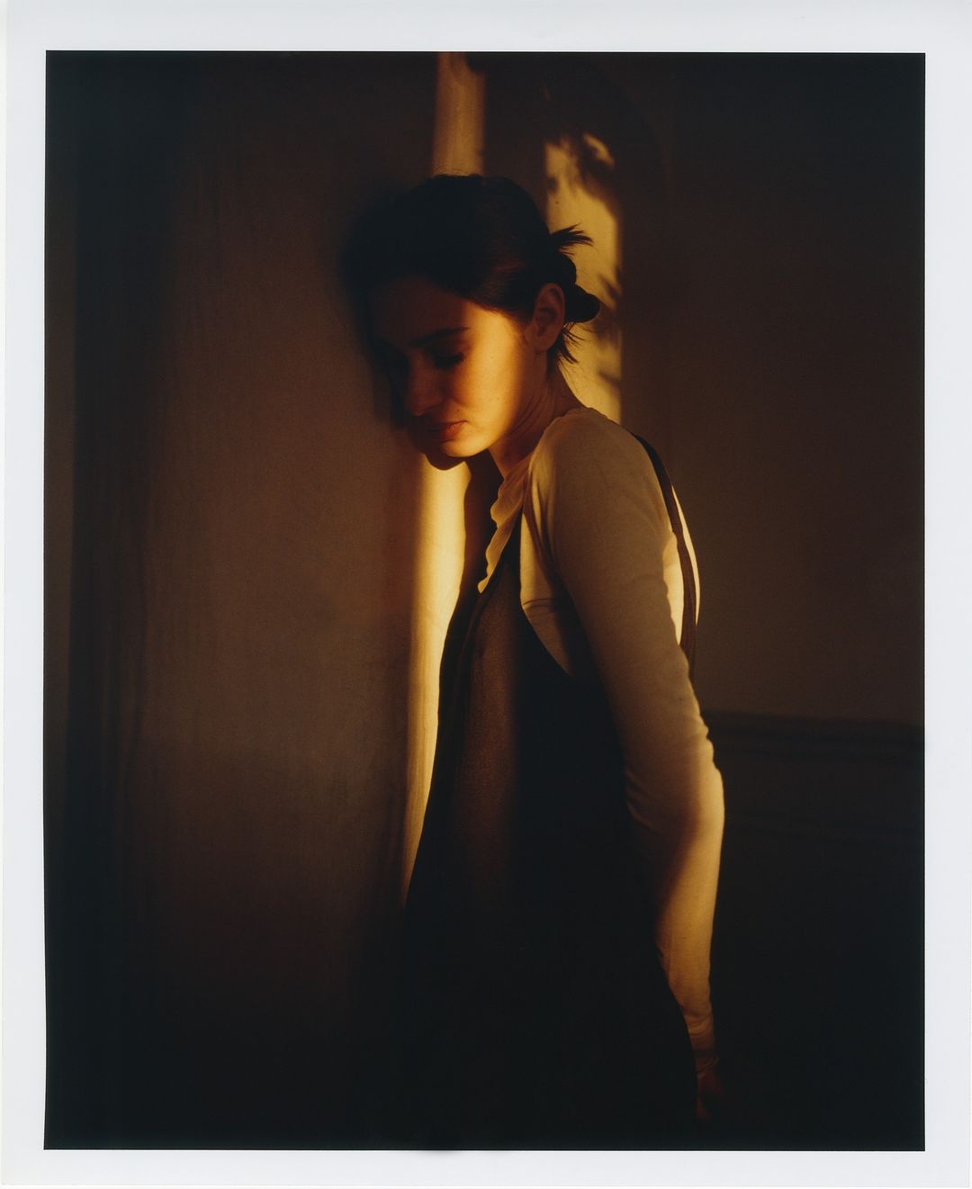 Woman stands near a wall, warmly lit with natural light accentuating her features and creating a soft, intimate atmosphere.