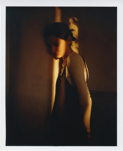 Woman stands near a wall, warmly lit with natural light accentuating her features and creating a soft, intimate atmosphere.