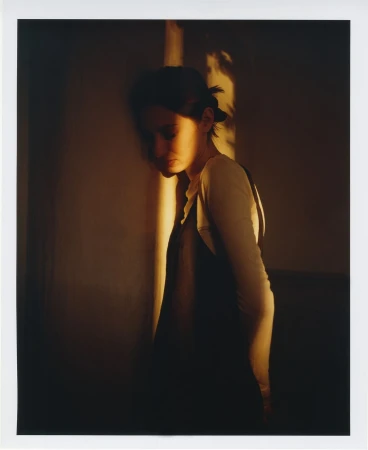 Woman stands near a wall, warmly lit with natural light accentuating her features and creating a soft, intimate atmosphere.