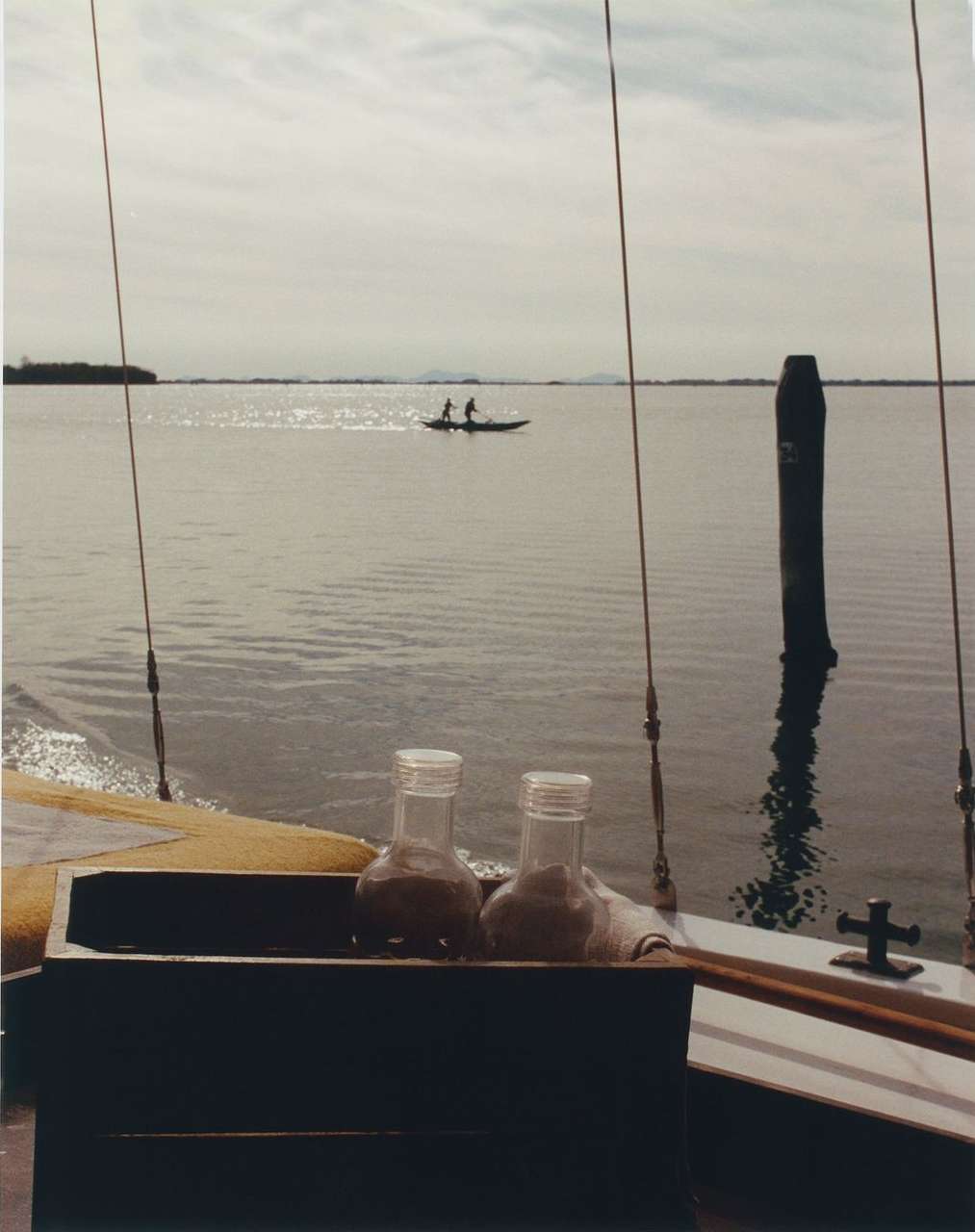 Two glass jars sit on a boat with two people in a canoe on the water visible in the distance under a cloudy sky during the daytime.