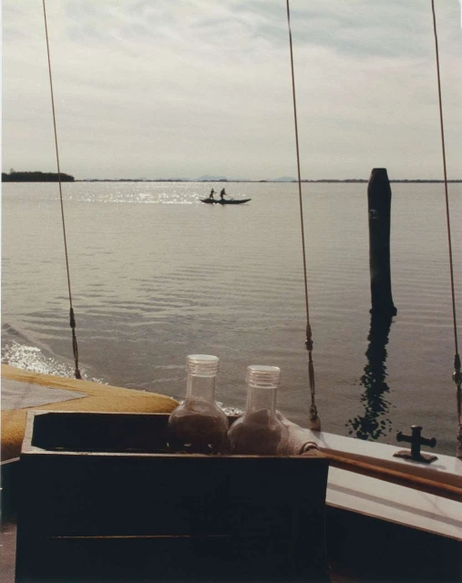 Two glass jars sit on a boat with two people in a canoe on the water visible in the distance under a cloudy sky during the daytime.