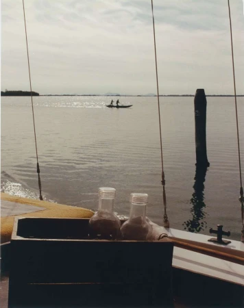 Two glass jars sit on a boat with two people in a canoe on the water visible in the distance under a cloudy sky during the daytime.