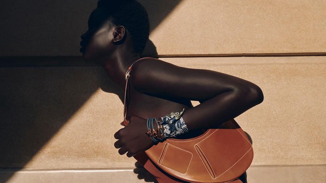 Fashion shot of a Black woman with a brown leather purse, accessorized with bracelets, against a tan wall with shadow patterns.