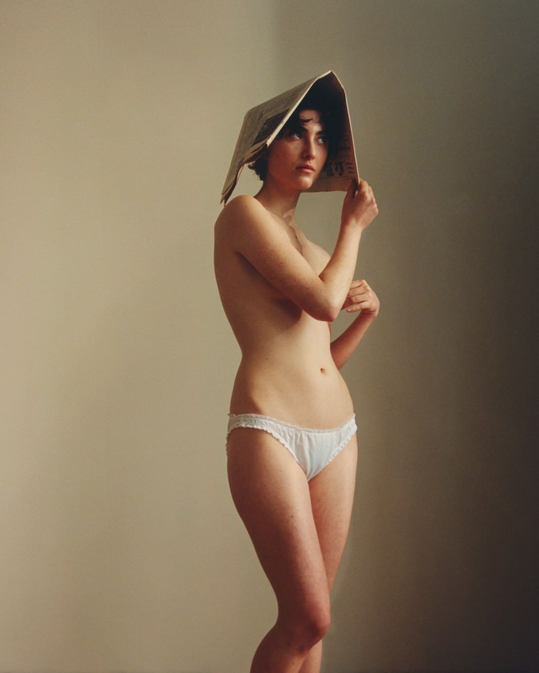 Nude woman in a photo studio with newspaper on her head and white underwear in front of a beige background with soft lighting.