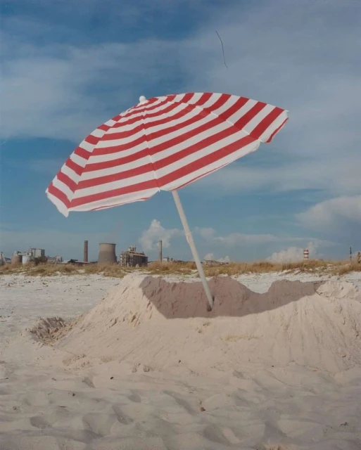Beach sandcastle under a striped beach umbrella, with a view of industrial buildings and chimneys on the horizon under a cloudy sky.