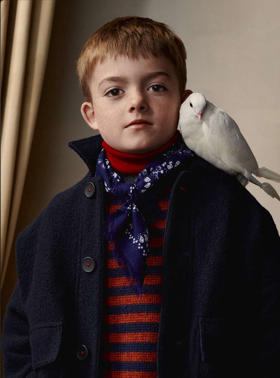 A boy in a navy coat poses indoors with a white bird perched on his shoulder, creating a timeless portrait.