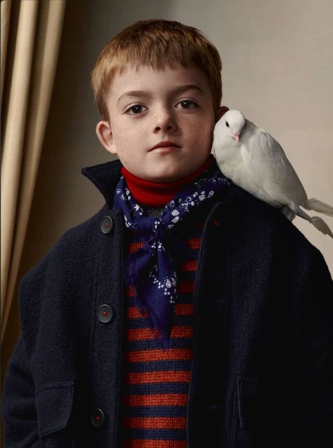 A boy in a navy coat poses indoors with a white bird perched on his shoulder, creating a timeless portrait.
