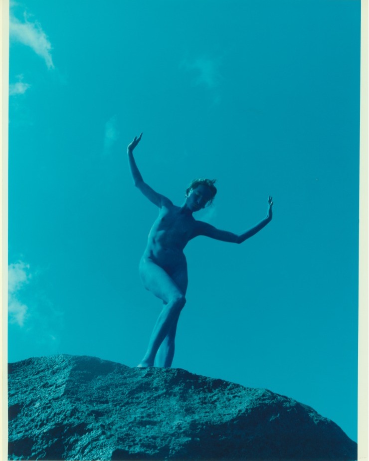 Artistic portrait of a nude adult female standing on a rock formation with arms raised against a vibrant blue sky with cloud accents.