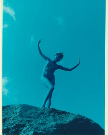Artistic portrait of a nude adult female standing on a rock formation with arms raised against a vibrant blue sky with cloud accents.