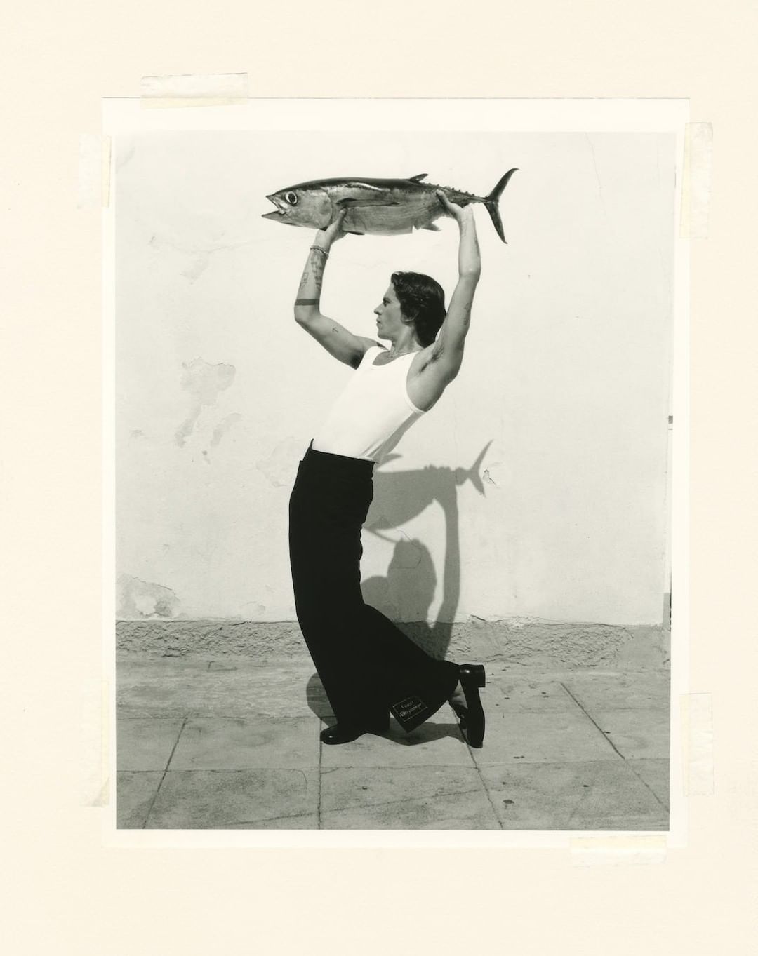 Black and white studio portrait of a person holding a large fish overhead while standing on a tiled floor against a plain wall.
