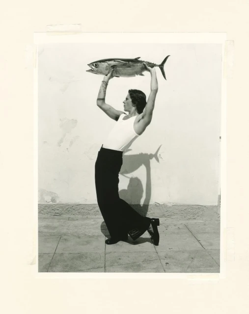 Black and white studio portrait of a person holding a large fish overhead while standing on a tiled floor against a plain wall.