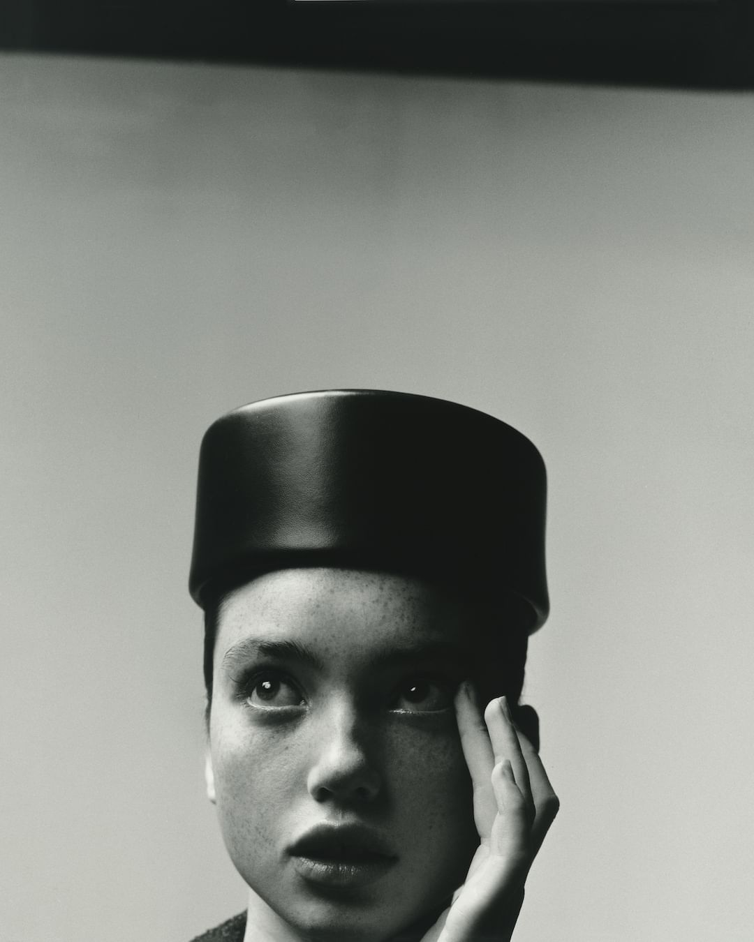 Close up portrait of a woman wearing a dark pillbox hat with her hand near her face against a plain background