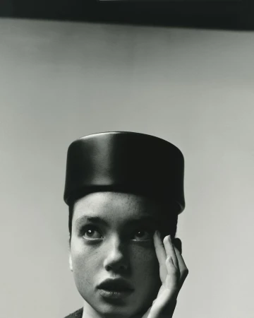 Close up portrait of a woman wearing a dark pillbox hat with her hand near her face against a plain background