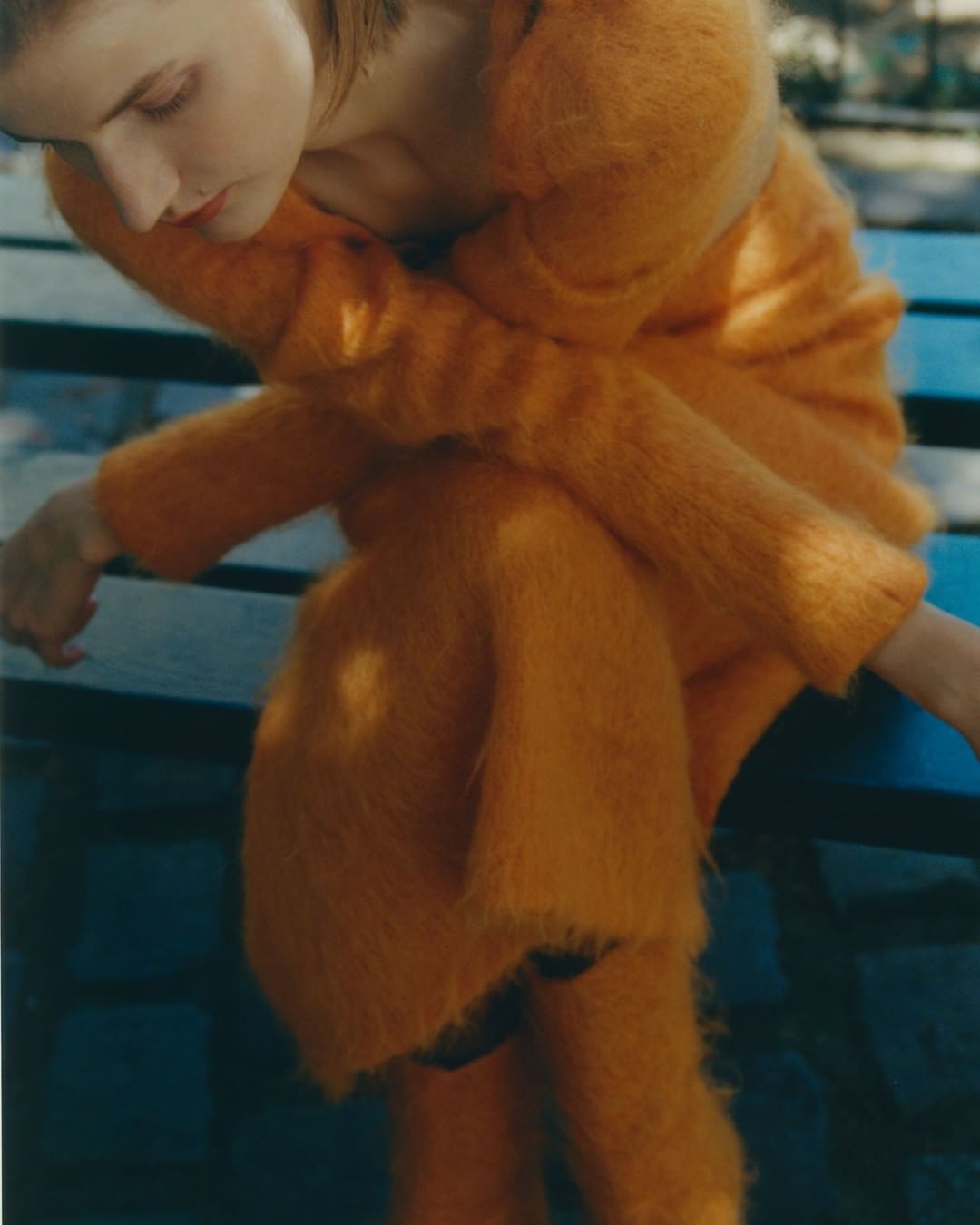 Woman in fuzzy orange sweater and skirt sits on a blue bench outdoors in soft light, gazing downward, fashion and style photography.