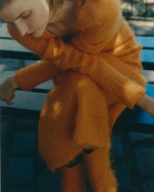 Woman in fuzzy orange sweater and skirt sits on a blue bench outdoors in soft light, gazing downward, fashion and style photography.