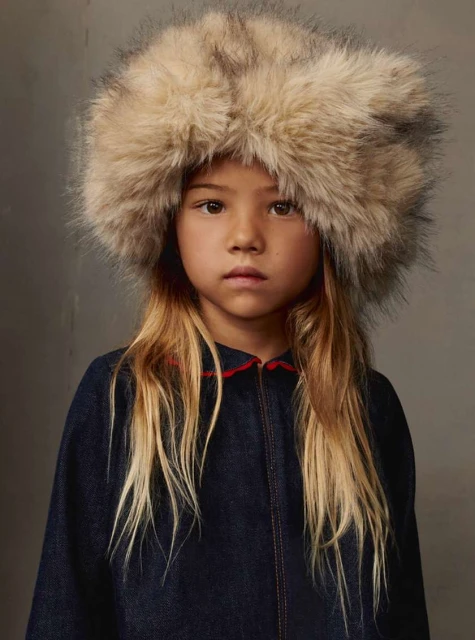Young girl wearing a large, furry hat and a dark blue denim-look jacket with a red collar, posing indoors against a neutral backdrop.