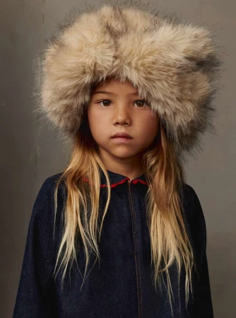 Young girl wearing a large, furry hat and a dark blue denim-look jacket with a red collar, posing indoors against a neutral backdrop.