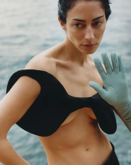 Woman in avant-garde black top poses with blue gloves against an ocean backdrop in a fashion editorial image.