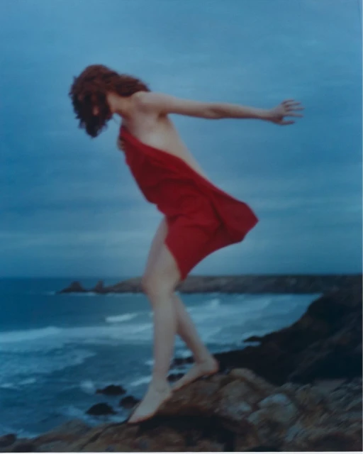 A woman in a red dress stands barefoot on a rock overlooking the ocean under a cloudy sky. Landscape orientation.