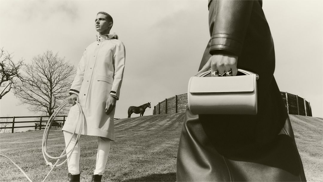 Two figures and a horse in a sepia-toned outdoor fashion shot. One figure holds a handbag, the other holds a rope in a field.