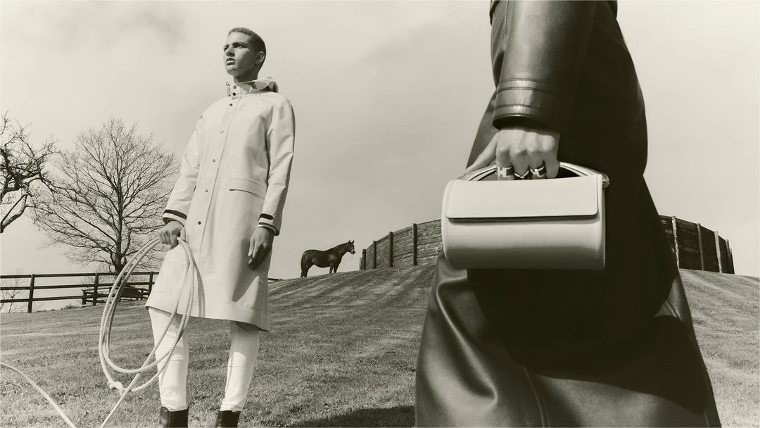 Two figures and a horse in a sepia-toned outdoor fashion shot. One figure holds a handbag, the other holds a rope in a field.