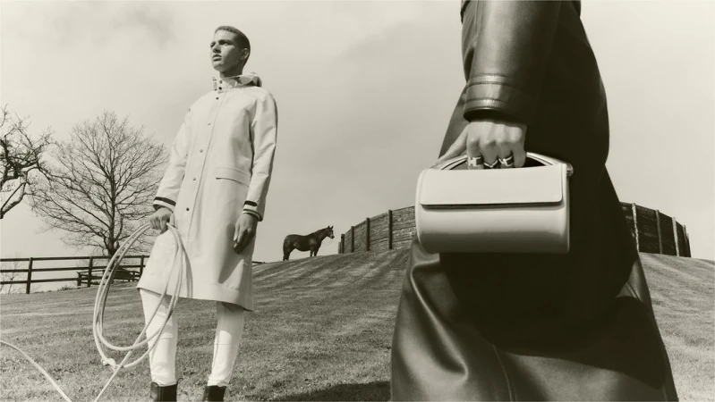 Two figures and a horse in a sepia-toned outdoor fashion shot. One figure holds a handbag, the other holds a rope in a field.