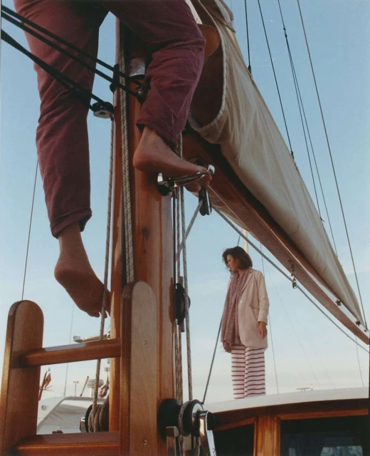 Woman in striped pants on sailboat watches person climbing mast in burgundy pants, set against a clear blue sky.