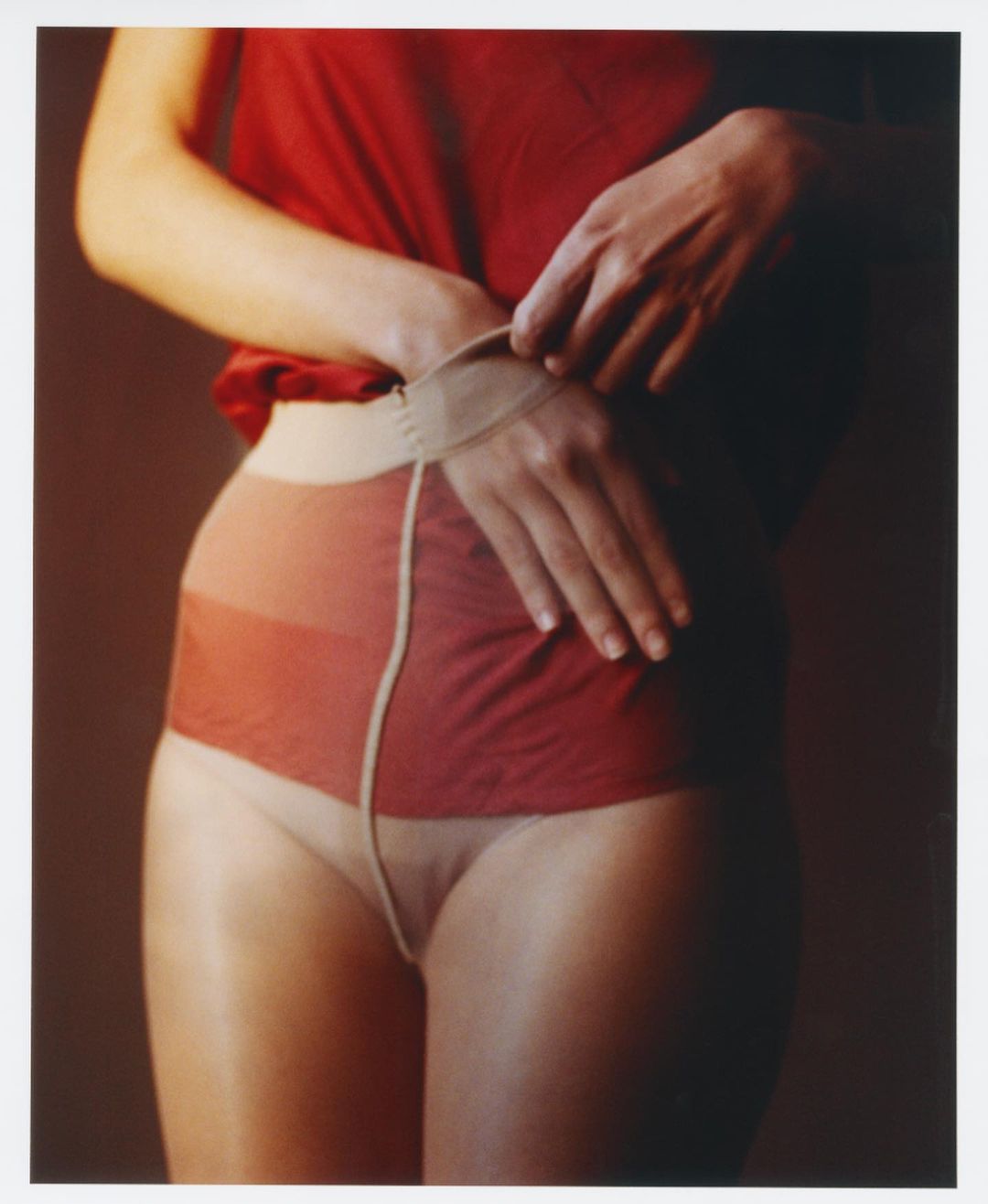 An adult female adjusts tan-colored tights over a red top, captured with soft studio lighting and a warm color palette in a medium shot.