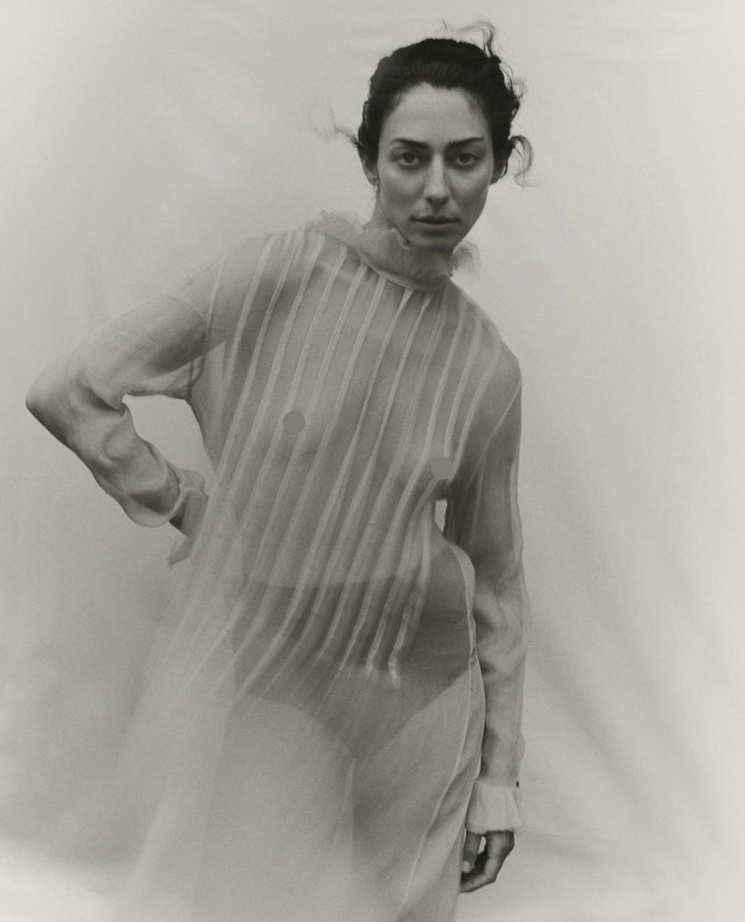 Adult woman in sheer dress poses in photo studio against a neutral background in a fashion photography style, monochromatic filter.
