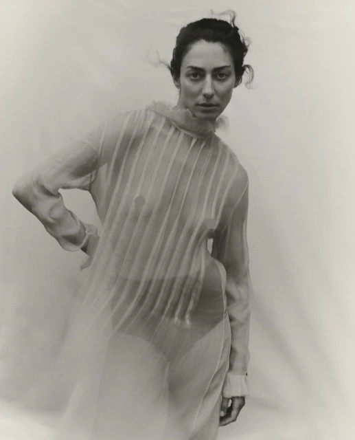 Adult woman in sheer dress poses in photo studio against a neutral background in a fashion photography style, monochromatic filter.
