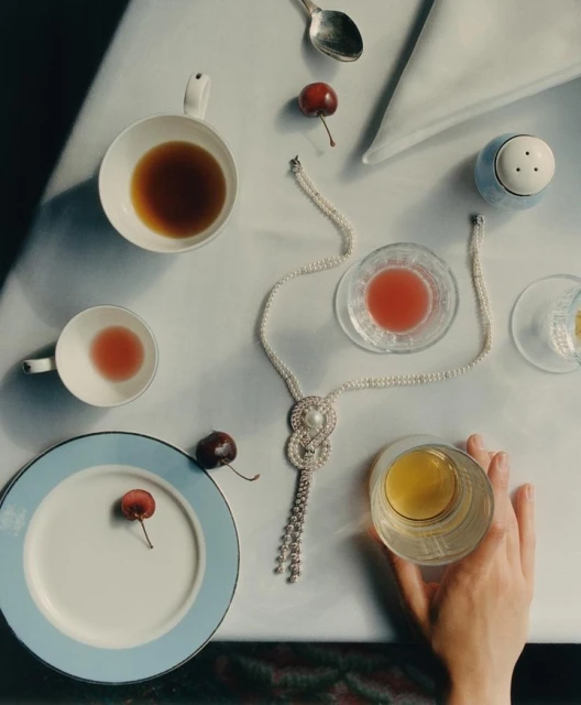 Overhead shot of a table setting with cups of tea, a pearl necklace, cherries, a plate, and a hand holding a glass of liquid.