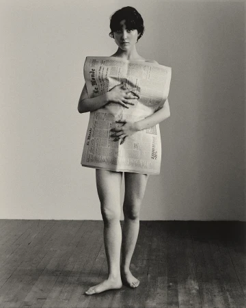Woman standing on wooden floor holding a broadsheet newspaper across her torso as a garment, captured in portrait orientation.