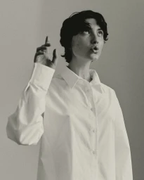 Portrait of a person with dark short hair wearing a white button-down shirt pointing upward against a seamless light background.