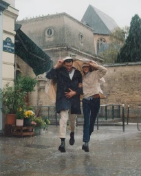 Man and woman laughing while running through heavy rain in a Parisian street, holding a large beige coat over their heads.
