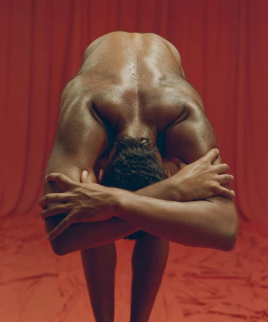Nude man with muscular physique bends forward, arms crossed, back facing the camera against a monochrome red draped background.