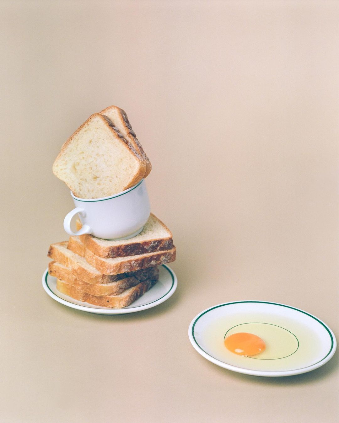 Still life with a stack of toast, a cup, and a raw egg on a white plate against a neutral background in soft, diffused lighting.