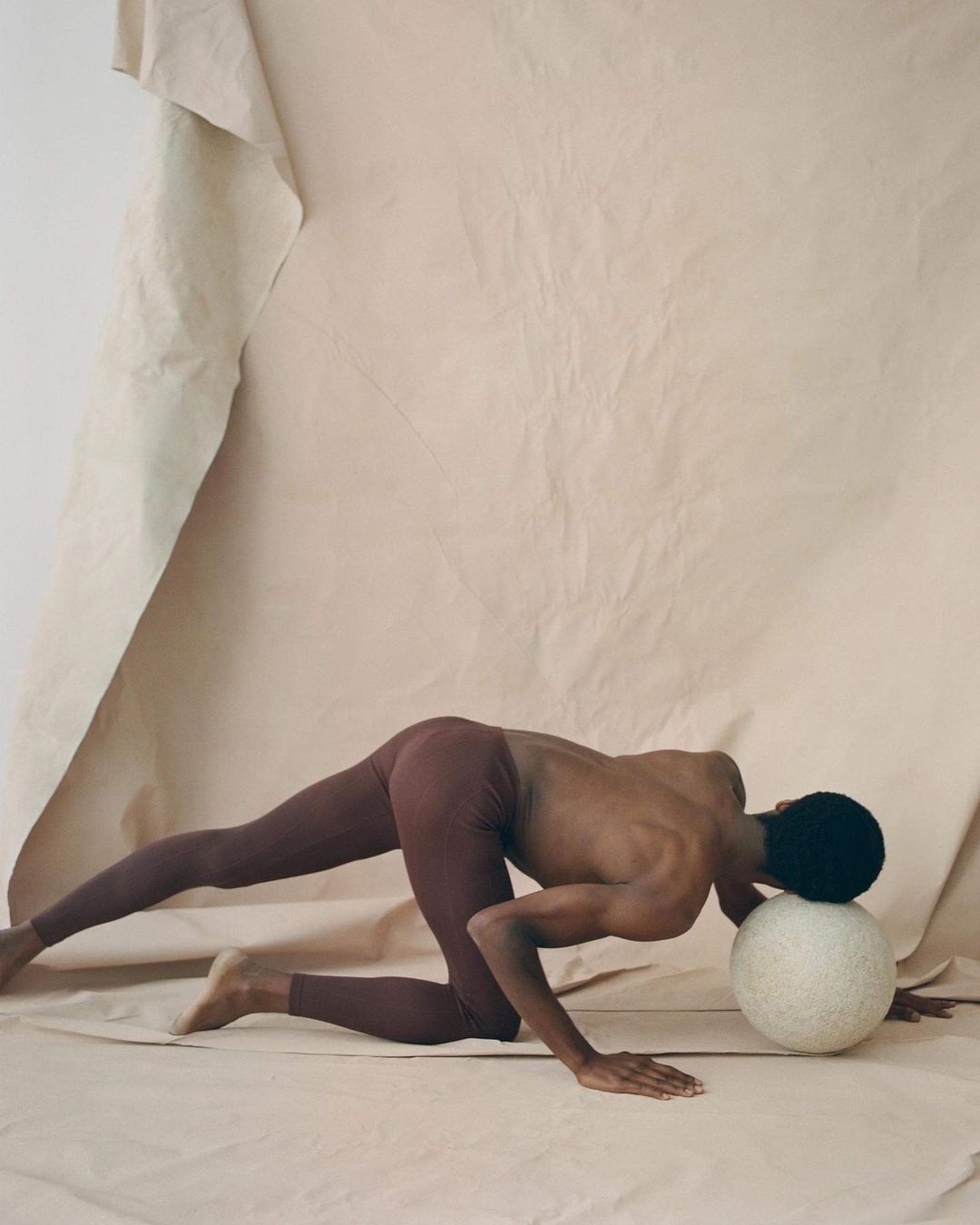 Adult male with black hair wearing leggings, leaning on white sphere in a neutral color studio setting with paper backdrop.