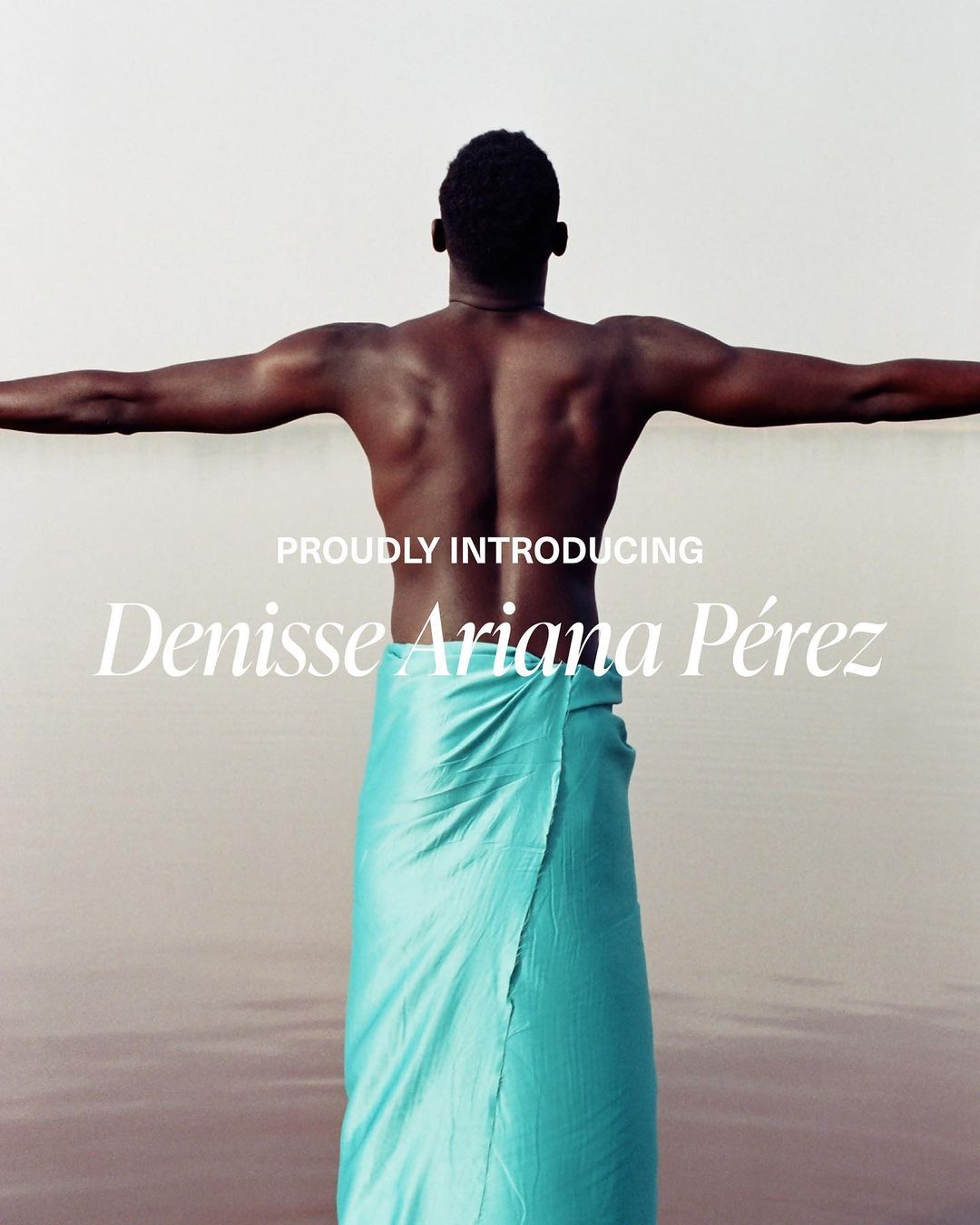 Back view of a dark-skinned man with arms outstretched wearing a turquoise wrap, standing in shallow, pink water under bright light.