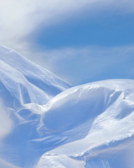 Abstract landscape of translucent material draped over formations under a blue sky with wispy clouds, creating an ethereal effect.