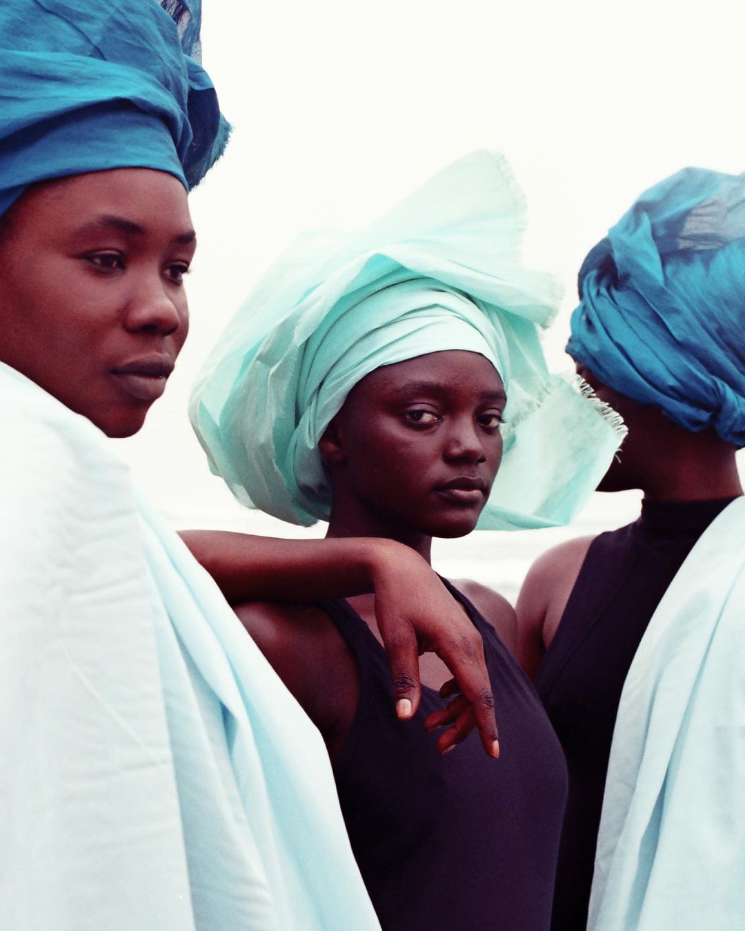 Three African women in blue and white headwraps pose outside against a neutral backdrop. Fashion, portrait photography.