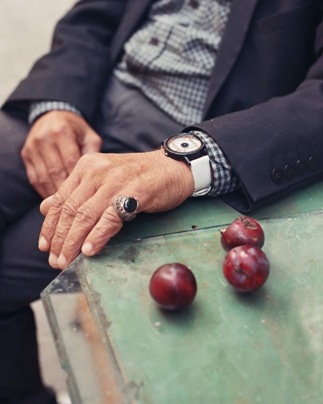 An elderly man in a suit wearing a ring and a watch sits at a table with plums. The light is natural and subdued.