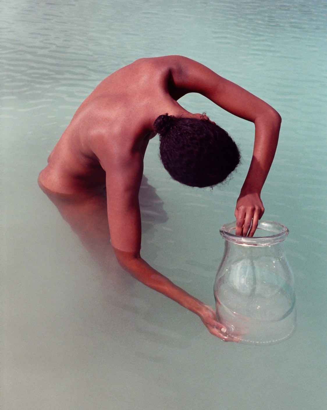 Woman in water holds clear vase, bending forward to submerge it, water reflecting soft light with ripples and visible skin texture.