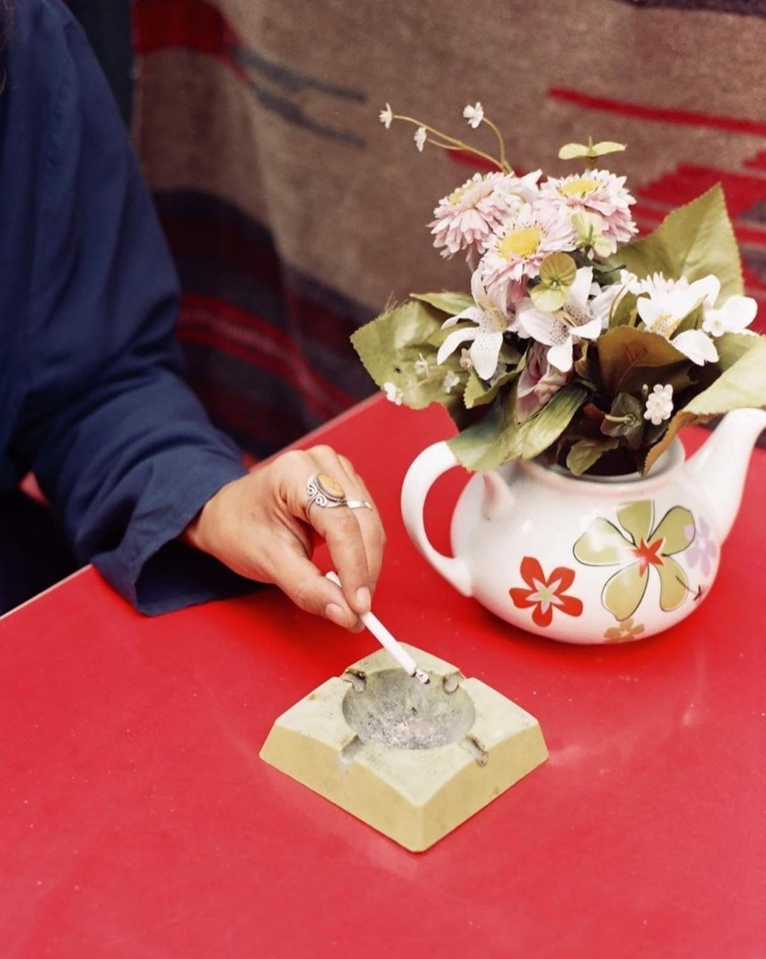 Woman with rings on her fingers at a red table putting out cigarette in ashtray next to floral arrangement in teapot.