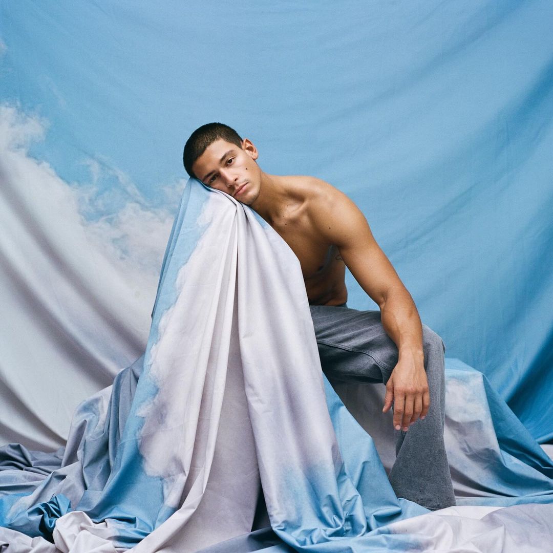 Man with bare chest, leaning on draped blue and white fabric, grey pants, neutral expression, studio lighting, against light blue background.