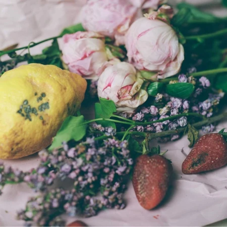 Still life arrangement of a large yellow lemon, fresh pink peonies, small purple flowers, and ripe strawberries on paper
